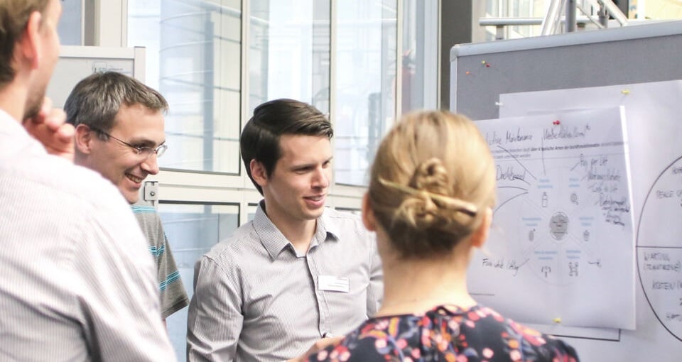 Four people stand in a group, discussing a diagram on a whiteboard in a bright, modern office setting. Two people face the board, while two others listen and engage in conversation. EFESO Management Consultants